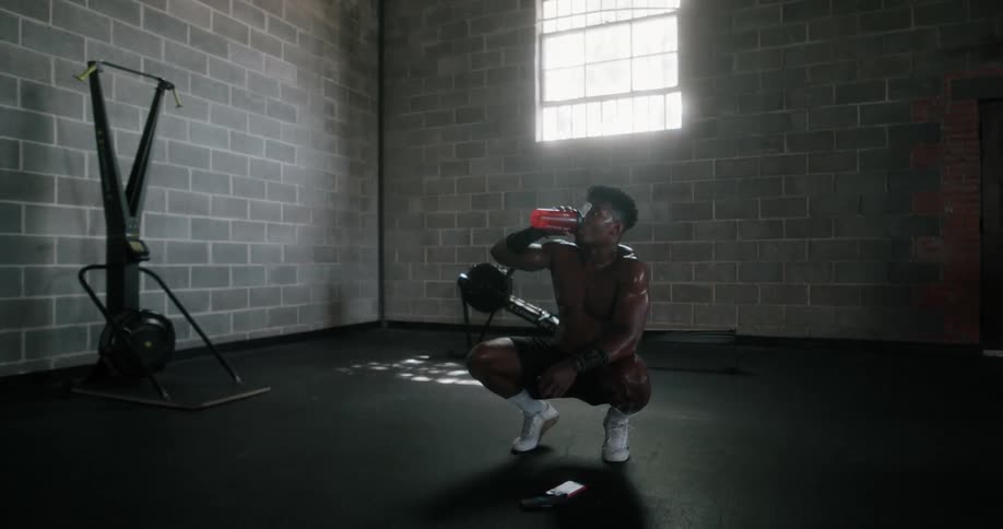A muscular and sweaty man squats while he recovers from exercising and drinks for hydration
