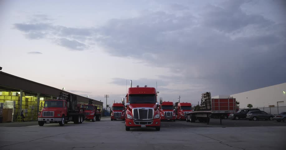 A fleet of red semi trucks sit in a parking lot outside a factory