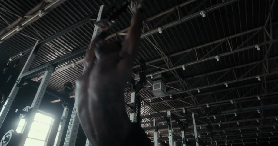 A shirtless man exercises on gymnastics rings hanging from the ceiling of a gym during the day