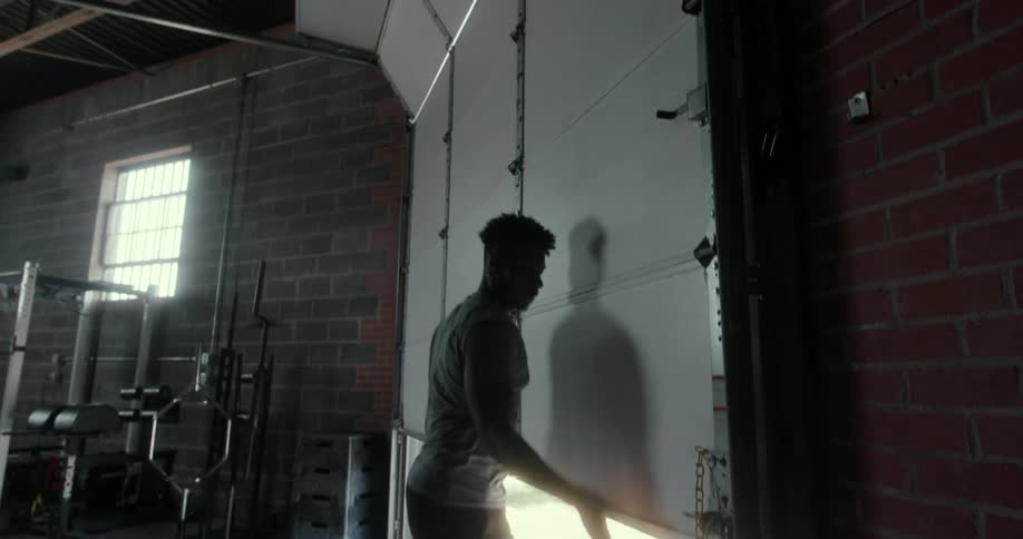 A muscular young man walks to a large door in a gym and opens it to let in light and fresh air