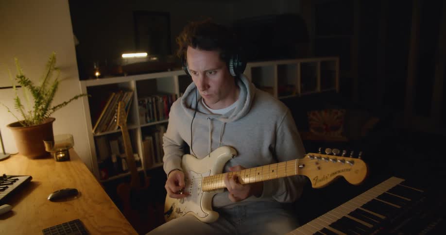 A man sitting at a desk plays a guitar while recording the sound with his computer