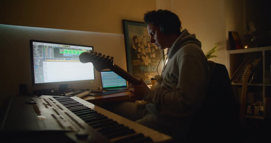 A young man sits in front of a computer and a keyboard and plays the guitar