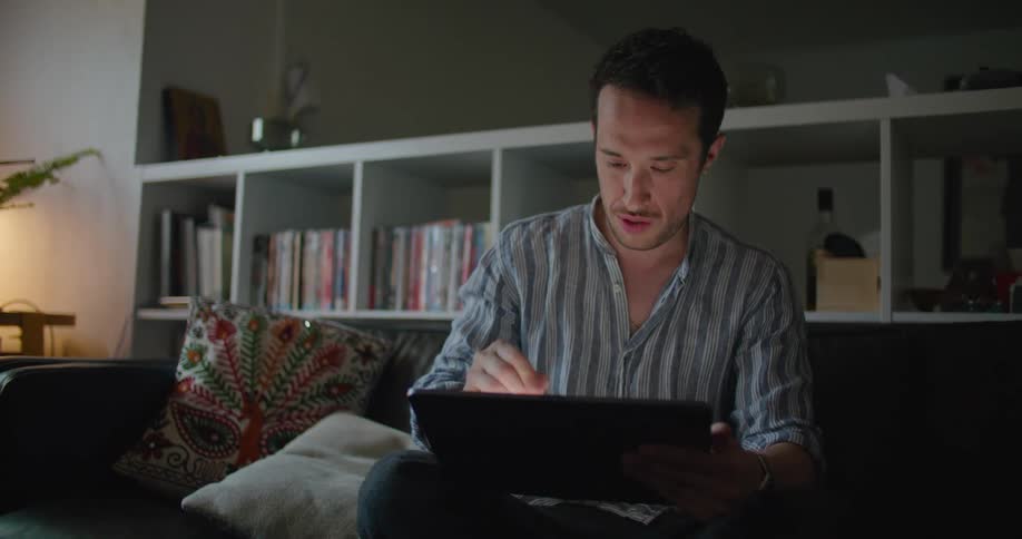 Man with grey shirt and jeans sits on black couch and uses stylus on tablet