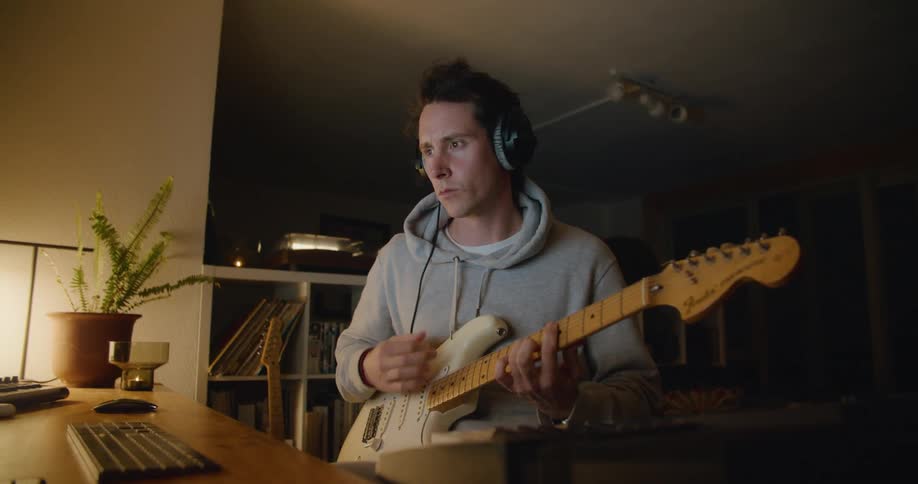 A male musicians sits at his desk when he picks up his guitar and starts strumming