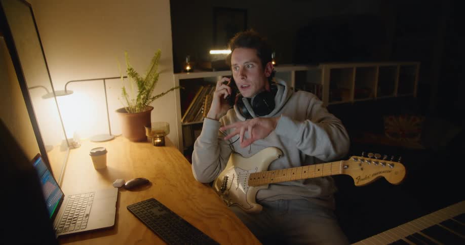 A man sits at a desk at home with his guitar in his lap talking on a phone and gesturing