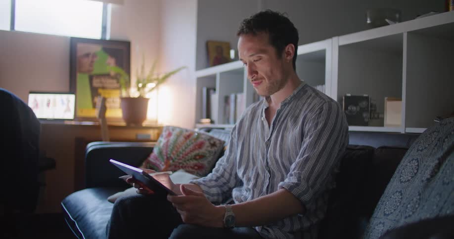 Man sits on sofa using laptop with laptop on desk in background