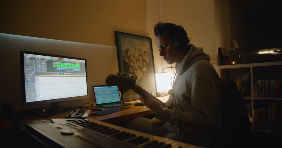 Man with headphones and gray hoodie plays bass guitar and looking at desktop