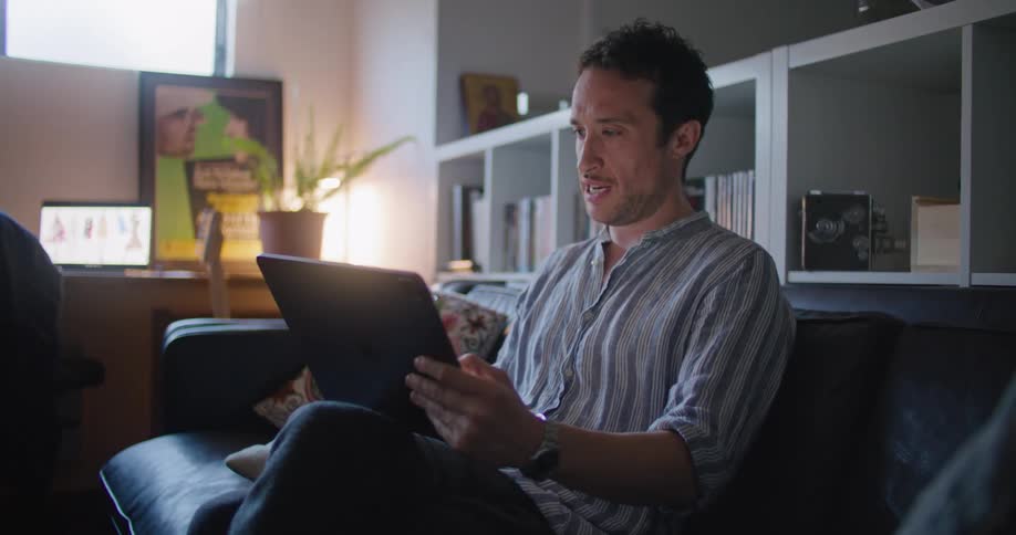 A man sits on his couch at home and holds a tablet that he video chats on