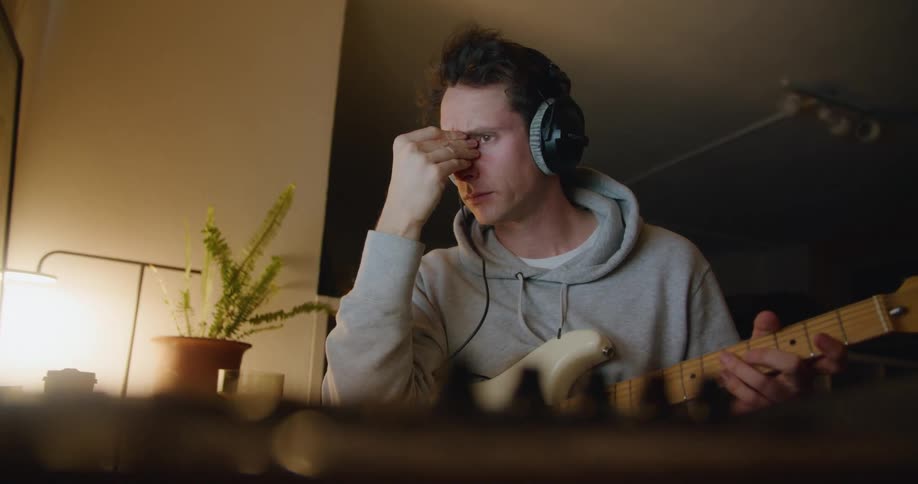A tired young man rubs his eyes while sitting at a desk holding a guitar in his hands