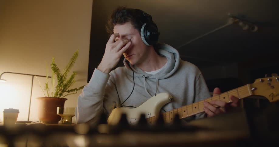 Musician holding guitar and wearing a headset interacts with laptop bobbing his head as he briefly rubs his eyes