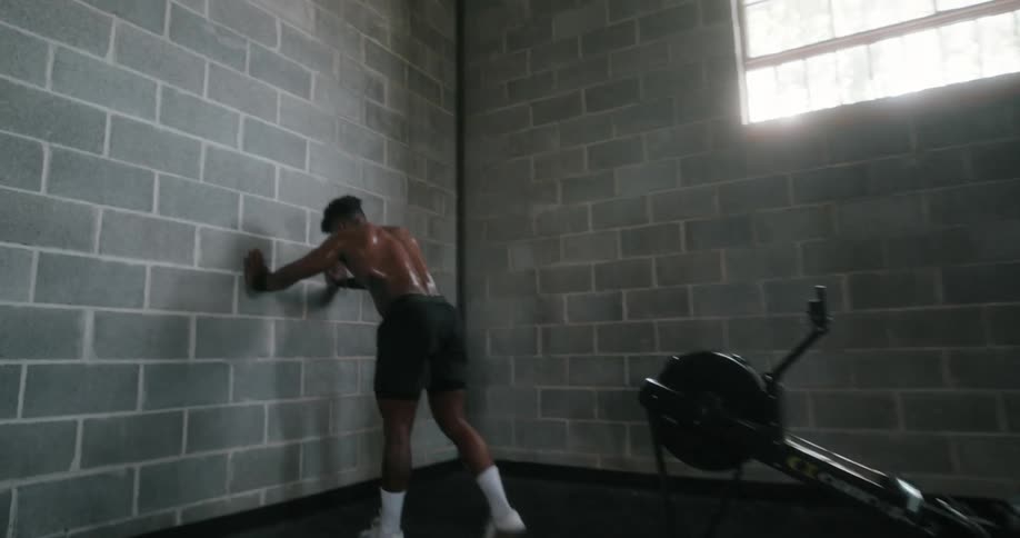 A man stretches his arm above his head and then leans against a wall and stretches his calf as he catches his breath after exercising