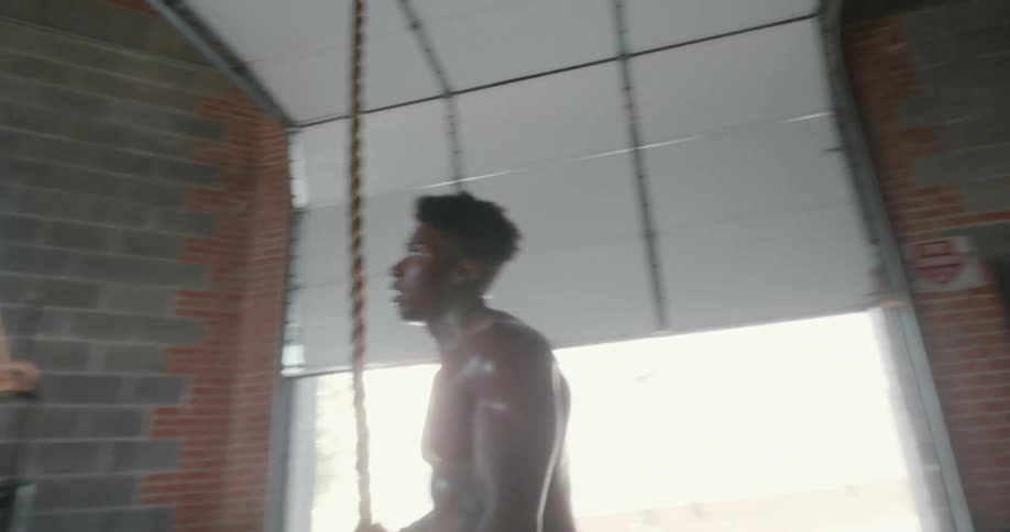 A muscular young man climbs down from a rope suspended from the ceiling above him inside of a gym space
