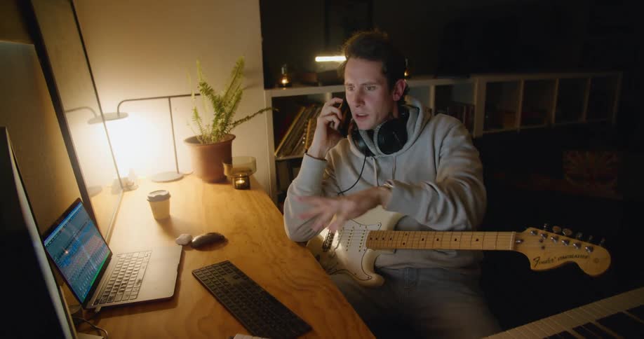 A man sits at a desk talking on a phone gesturing with his hands while holding a guitar in his lap
