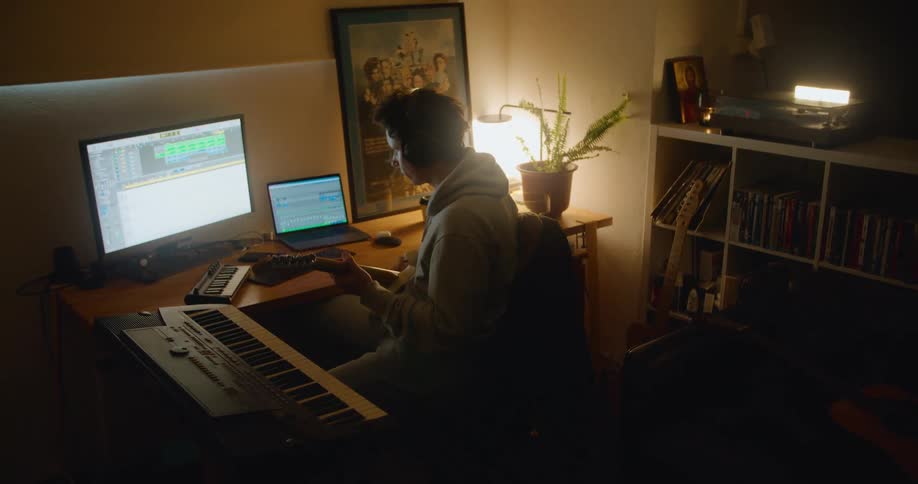 A man wearing headphones while sitting at a desk plays a guitar while recording the sound with audio software on a computer