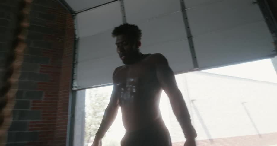 Sweating bare chested walks to open door and back before climbing rope in gym