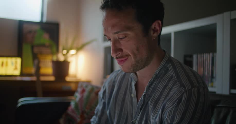 A man sits at home and smiles as he draws on a small white board he is holding