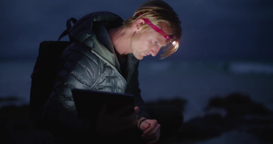 Man crouches on beach using tablet as he examines area with waves rolling in background at night