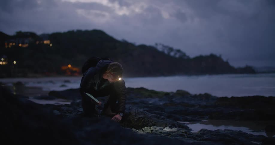 A man wearing a light on his forehead and holding a tablet walks on a beach and crouches down to grab something