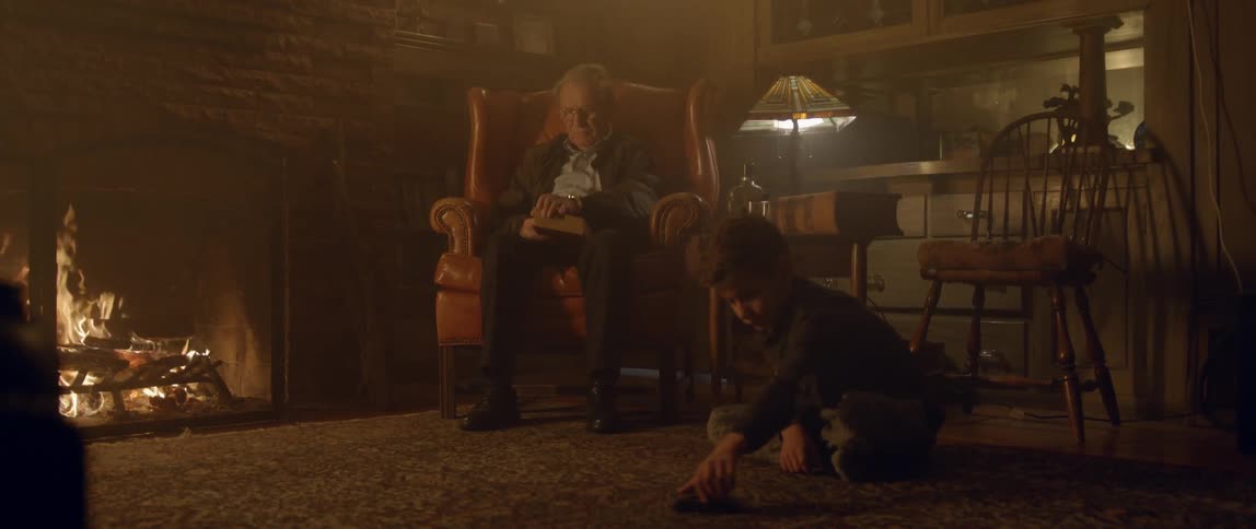 An old man sits in a chair reading a book while a young boy plays with a car on the ground near him