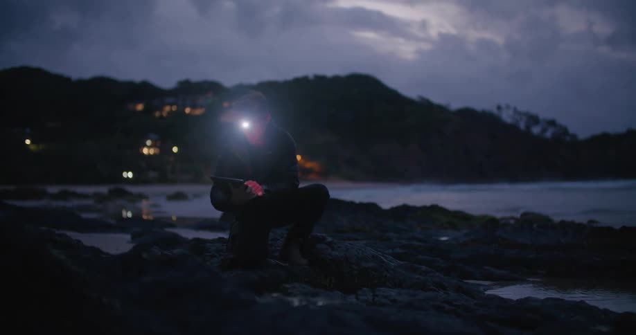 Man wearing headlamp crouches on beach examining ground and making notes in tablet before looking around