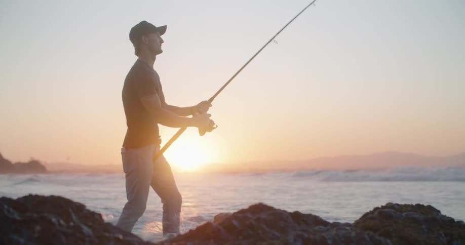 A man stands on the shoreline and reels his fishing rod in