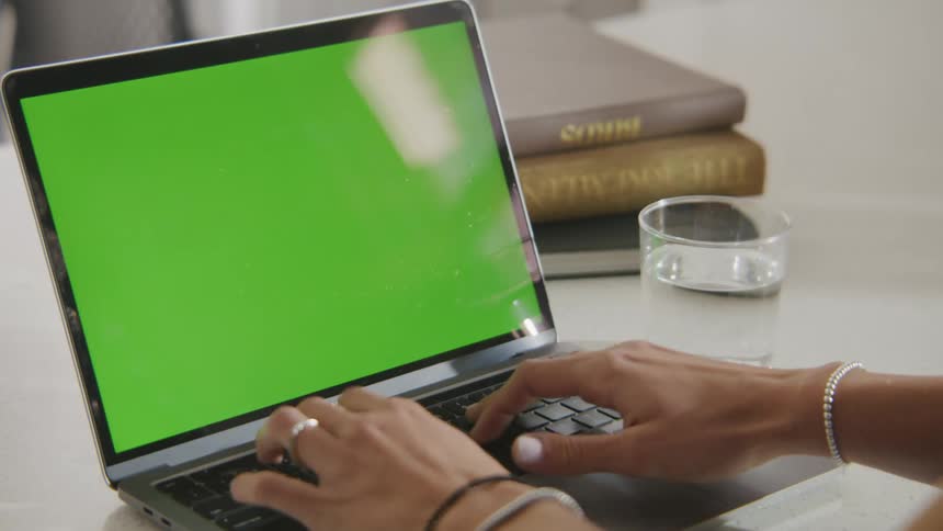 Woman types on laptop placed near glass of water and hardback books