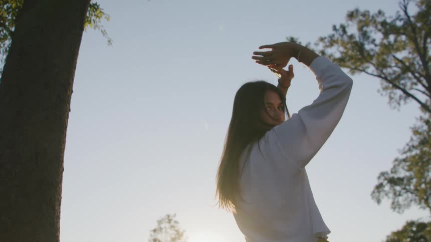 Woman wearing a white shirt is standing next to a tree with her arms raised over her head