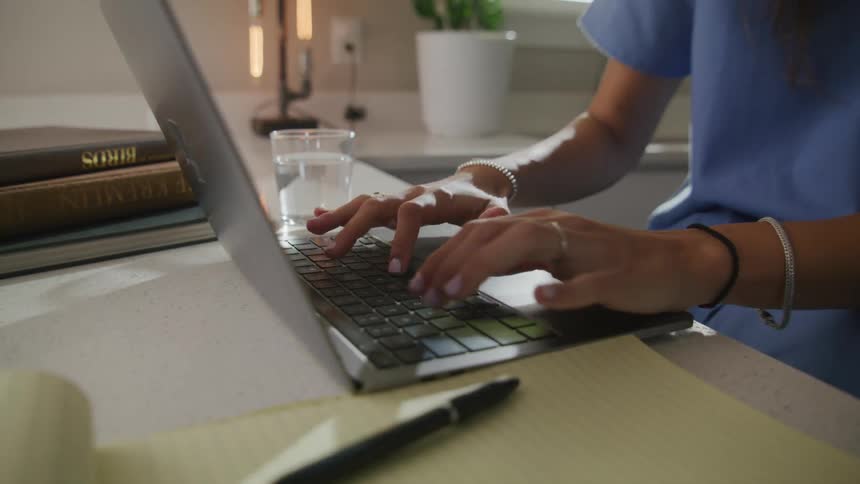 A woman in a nurses uniform smiles while using her laptop in the kitchen