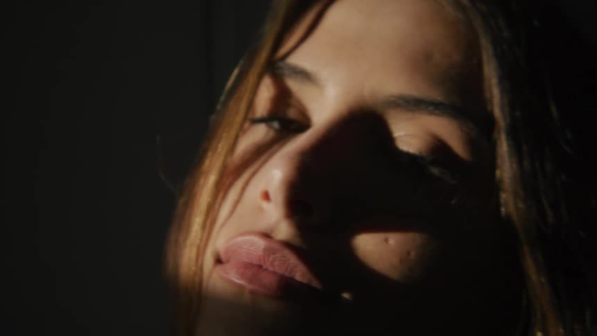 A young woman in a dark room smiles into the camera while sunlight illuminates her face