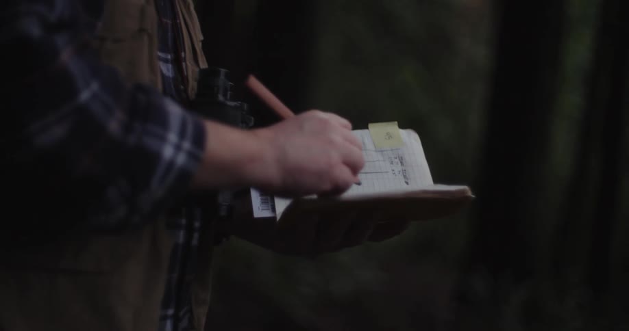 Man wearing vest writes in notebook before gazing through binoculars