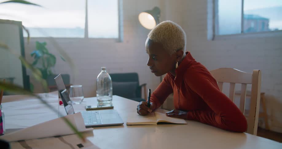 A young woman sitting at a table writes in a notebook while using a laptop to participate in a video conference