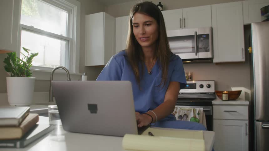 Young nurse sitting in the kitchen at home typing on a laptop