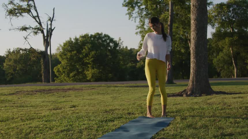 A young woman walks to a mat in a grassy field and steps on to it to exercise