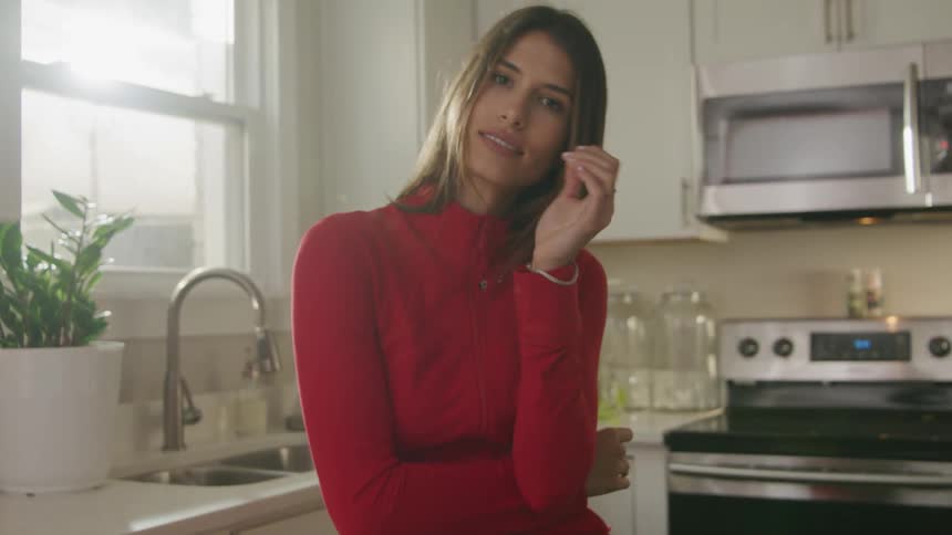 A woman wearing a red turtleneck sweater is standing in a kitchen and rests her head on her hand