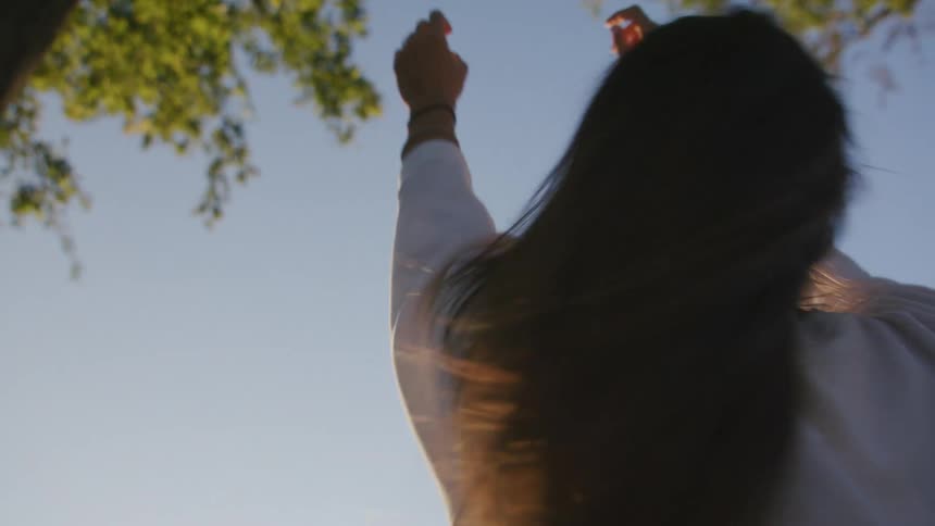 A woman raises her arms up and dances under tall trees