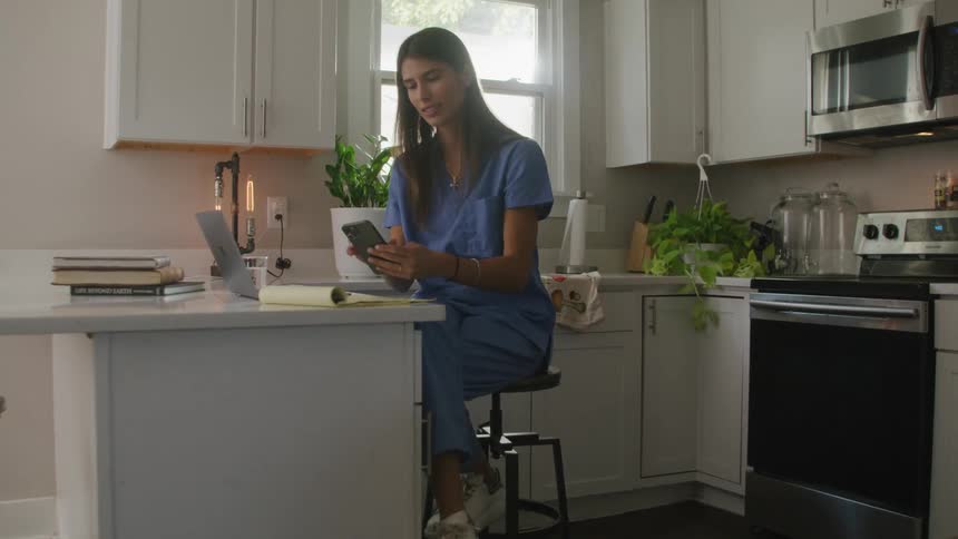 A woman is sitting at a kitchen counter with a computer and notepad when she answers her phone and stands up and turns away