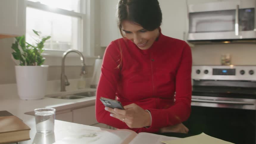 A woman sits in her kitchen and picks up her cell phone and laughs as she looks at it