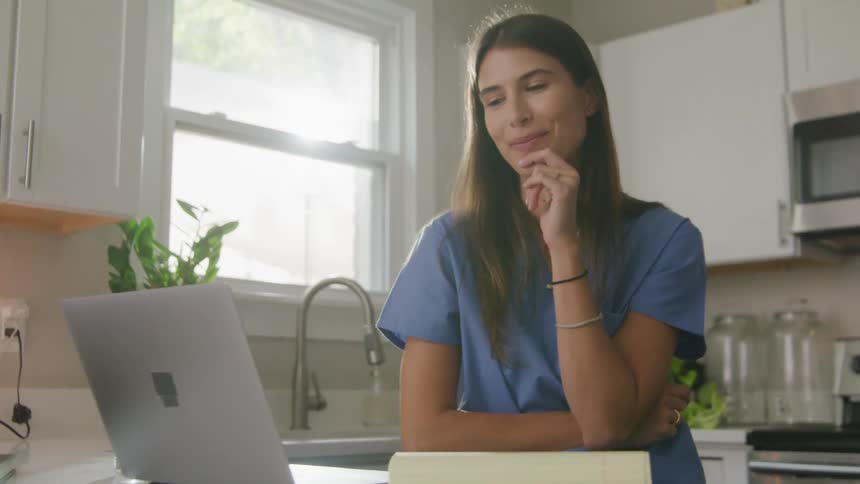 Woman with brown hair and blue shirt makes video call on grey laptop in the kitchen smiling