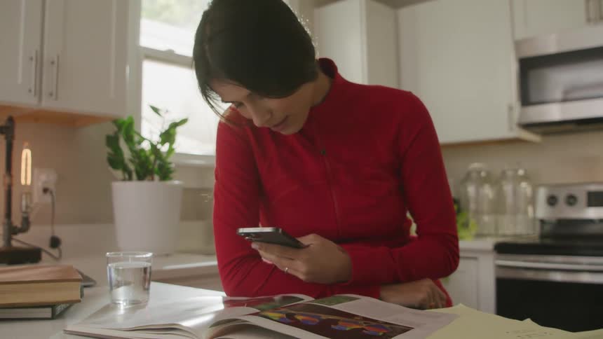 A woman leaning on her kitchen counter looking at her phone with a book and notebook open in front of her