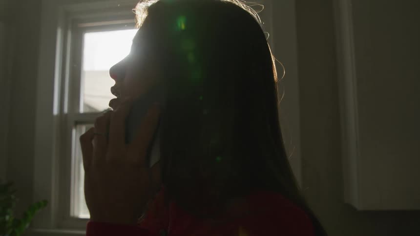 A woman talking on the phone in front of a sunny window indoors