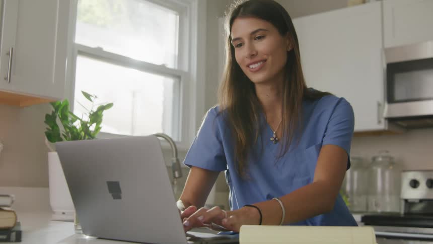 Woman wearing scrubs talks and smiles while standing in kitchen typing on laptop