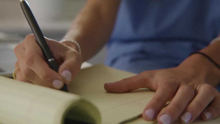 A woman wearing blue is sitting at a counter and writing on a tablet of yellow paper