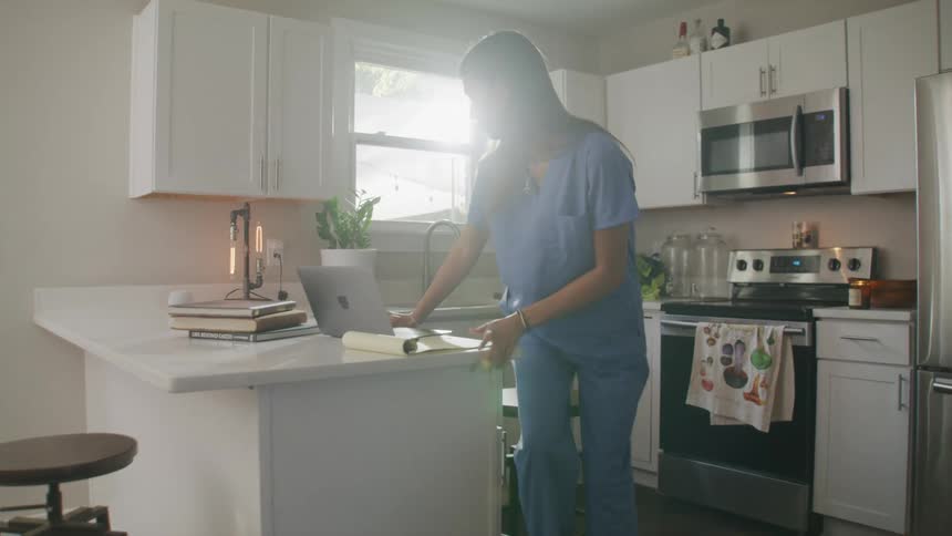 A woman wearing blue carries a laptop computer to a kitchen counter and sits down