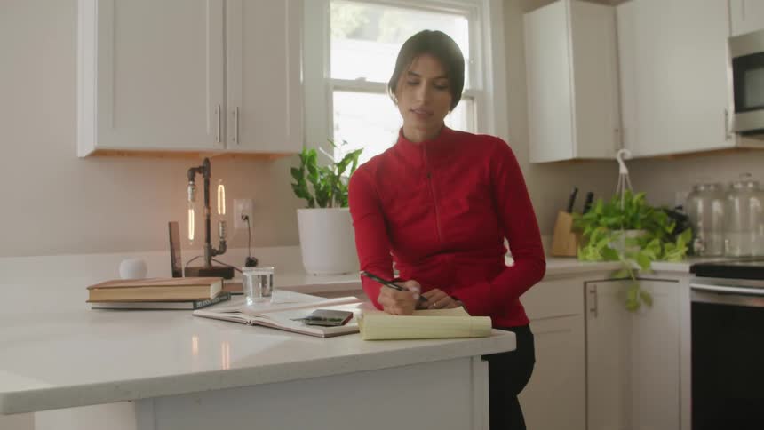 Young woman wearing red sweater writing on a notepad in the kitchen at morning