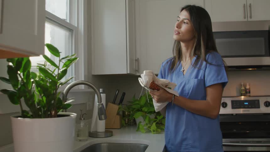 A woman in a nurses uniform wipes her hands while looking out her kitchen window