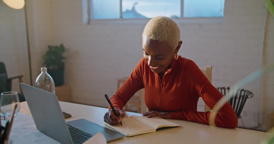 A young woman writes in a notebook while participating in a video conference using a laptop