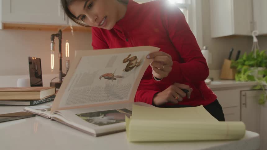 A woman wearing a red shirt is standing at a kitchen counter looking at a book with a notepad next to her