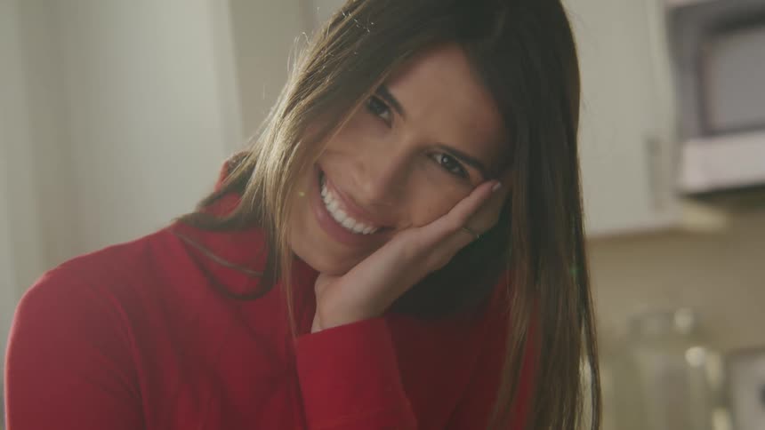 A woman wearing a red turtleneck sweater is standing in a kitchen with a big smile on her face as she rests her head on her hand