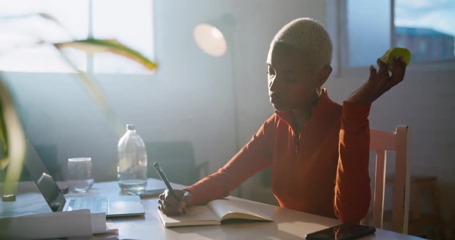 A woman in a room sitting at a table and using a laptop with a wall and window in the background