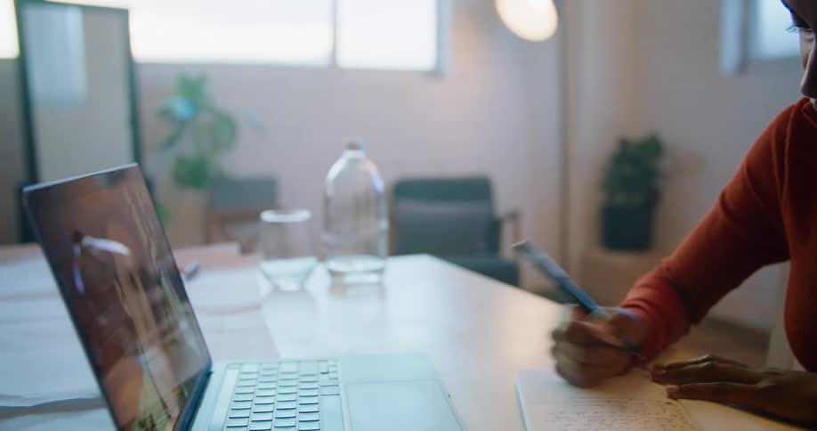 Woman with short hair earrings and orange zip up makes video call on laptop and takes notes in the office